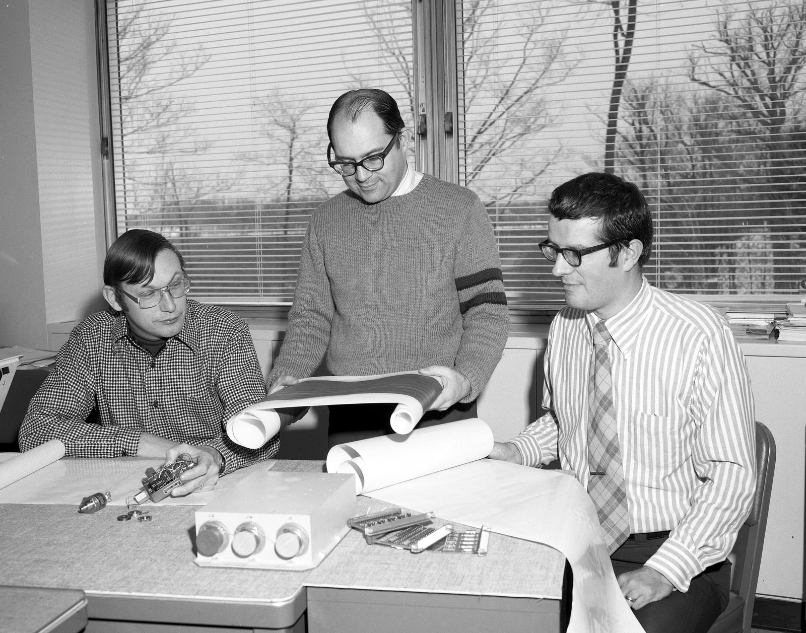 Tres ingenieros revisan juntos un listado impreso de computadora sobre un escritorio, 1976