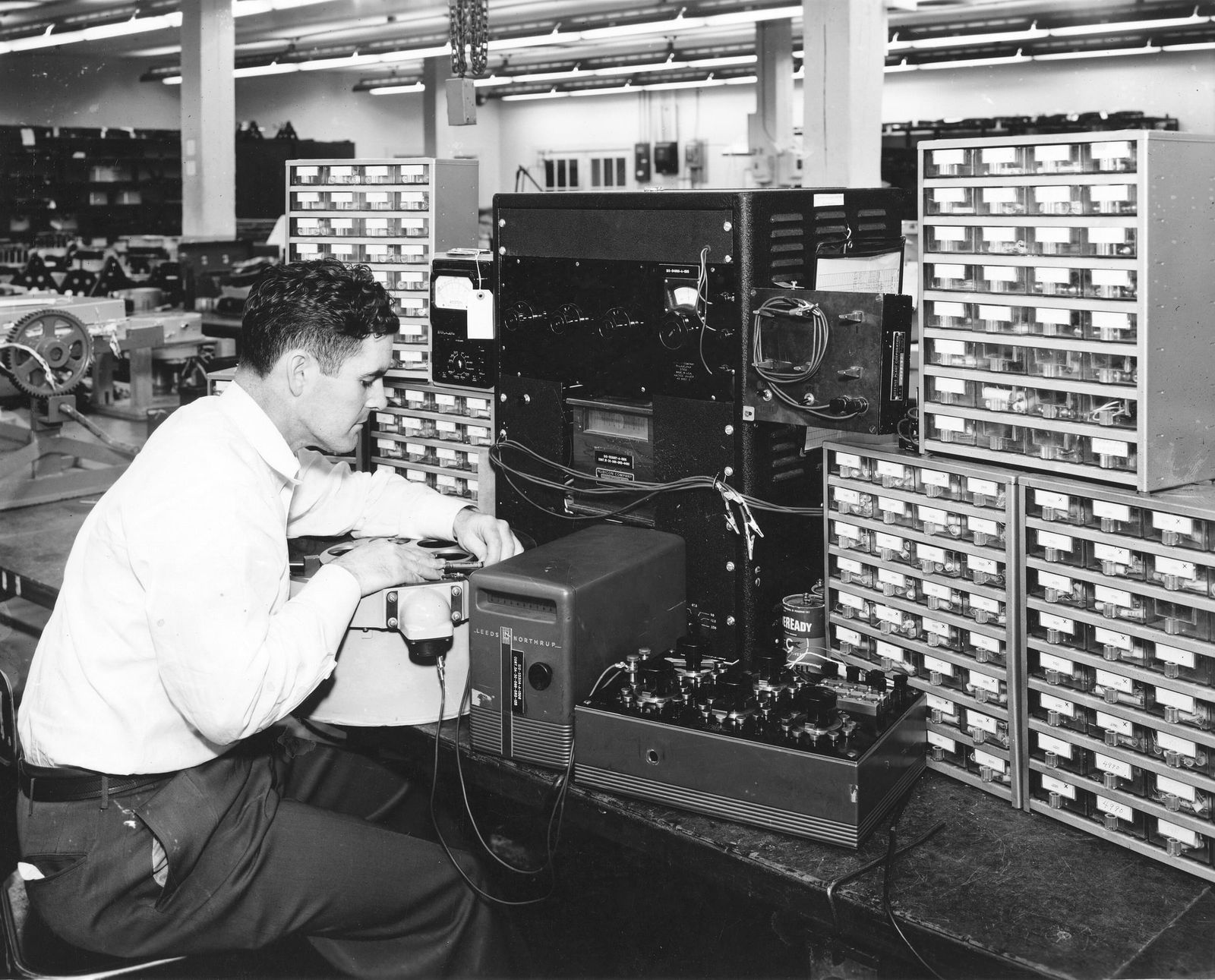 Técnico probando componentes eléctricos rodeado de estanterías de piezas, 1953